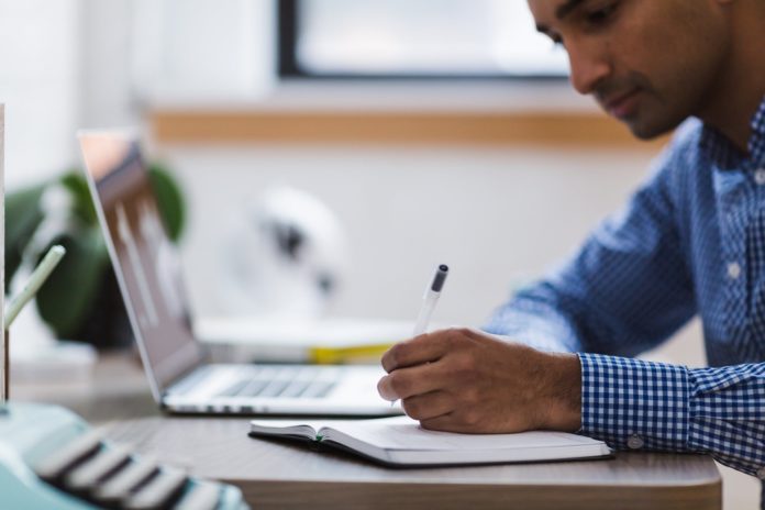 A employee takes notes at their desk beside a laptop.